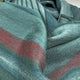 Pallalanga baby alpaca wool blanket – a green wool blanket patterned with red and blue stripes is seen draped and folded. The blanket's texture and colors are prominent, resting in an indoor setting