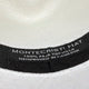A close-up of a hat's interior label reads: MONTECRISTI HAT 100% PAJA TOQUILLA HANDWOVEN IN ECUADOR on a textured white fabric background within a black band.