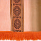 The textile features a vertical geometric pattern in the center, with horizontal orange fringes at the bottom. It is set against a gradient background of peach and beige stripes.