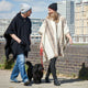 Two people wearing ponchos and beanies walk a black fluffy dog on a city sidewalk, with tall buildings visible in the background under a clear sky.