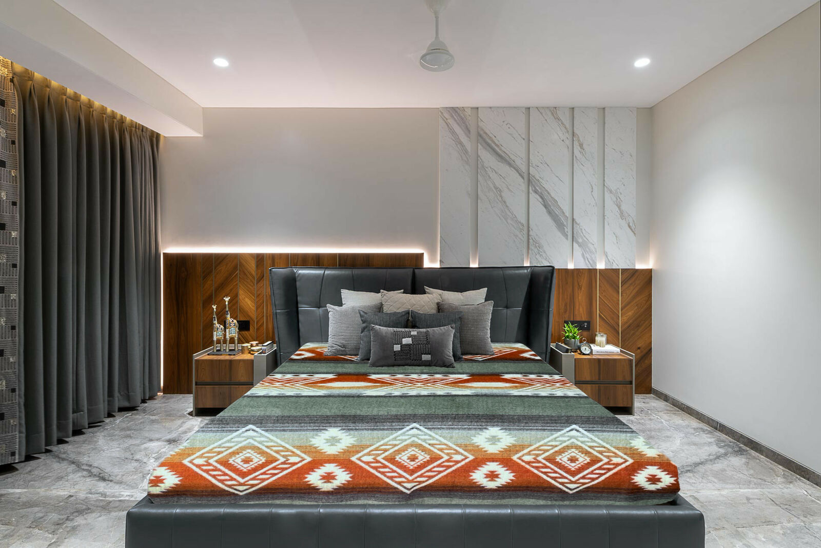 A bed with geometric-patterned bedding and numerous pillows is centered against a backdrop of wooden and marble panels. It is flanked by nightstands with decor, curtains, and soft lighting.