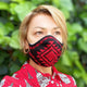 A person wears a patterned black and red face mask and a red shirt with architectural designs, standing against a leafy green background.