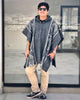 A person stands smiling, wearing sunglasses and a patterned poncho, on a tiled balcony overlooking a blurred cityscape through a windowed wall.