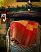 Yacupungu baby alpaca wool blanket – a striped, red-orange blanket drapes over a woven chair in a cozy room, with a Christmas stocking and green garland adorning a stone fireplace mantle