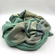 Saquisili baby alpaca wool blanket – a soft, multi-striped green blanket is loosely crumpled, sitting on a plain white surface, showcasing its textures and folds