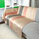 Tilivi baby alpaca wool blanket – a multicolored, striped throw blanket drapes over a gray sofa. The setting includes a white shelving unit displaying decorative items and a geometric-patterned blanket on the floor