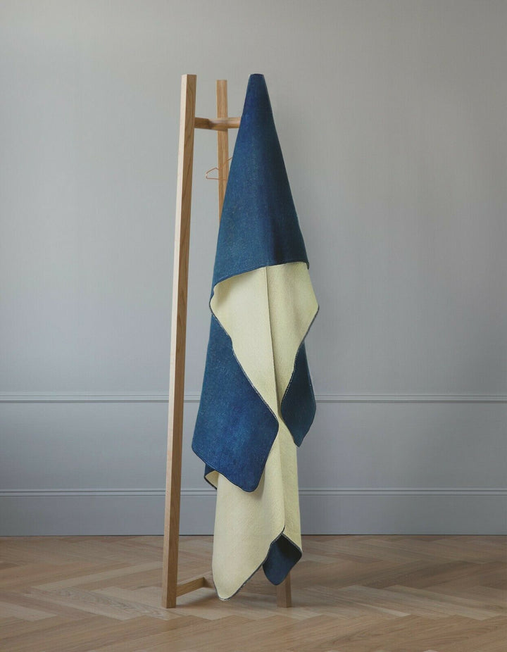 A folded, two-toned blue and cream blanket hangs on a minimalist wooden stand, situated against a light gray wall on a wooden floor.