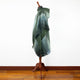 A green shawl drapes over a wooden mannequin stand, set against a white wall and hardwood floor, showcasing gradient fabric with fringed edges.