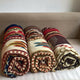Three rolled blankets display vibrant geometric patterns and earthy tones. They rest side by side on a beige sofa, set against a plain white wall backdrop.