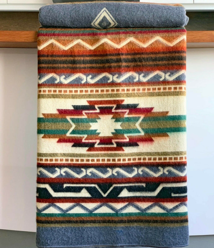 A colorful blanket hangs over a wooden bar, featuring geometric patterns in red, green, beige, and blue. The background is a simple indoor setting, with white cabinetry.