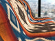 Qhusi extra large alpaca wool blanket – a patterned textile with bold, geometric designs in blue, orange, and white drapes over a surface near a window, with a blurred cityscape visible in the background