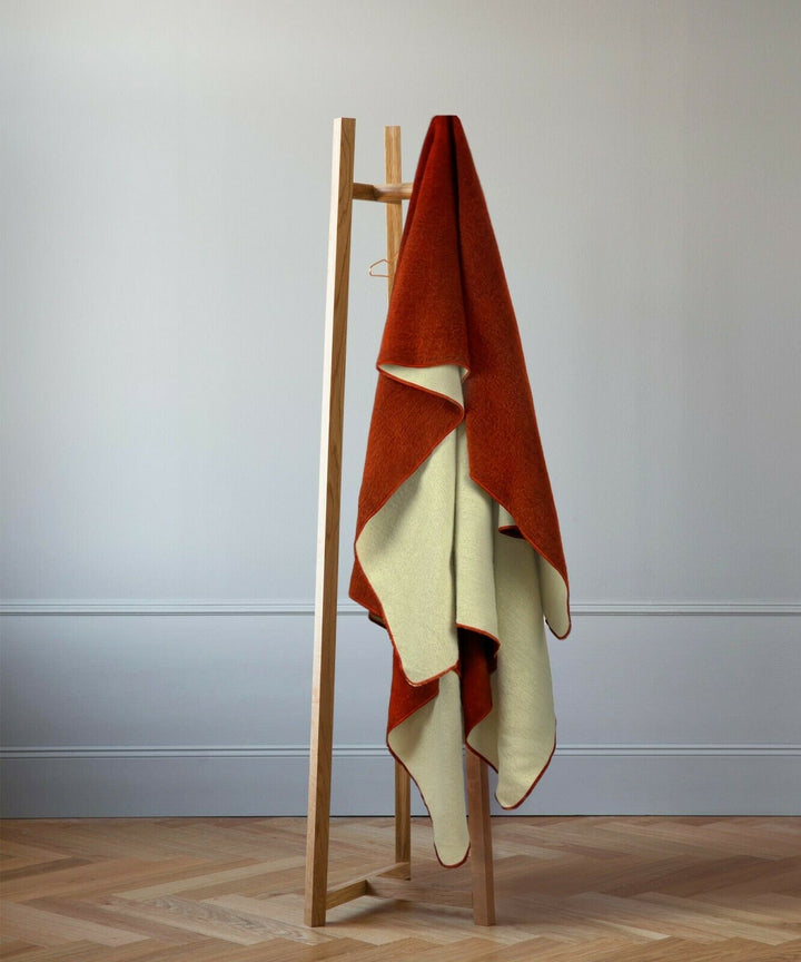 A wooden stand holds a red and cream blanket, draped neatly. The scene is set against a minimalist room with pale walls and herringbone-patterned wooden flooring.