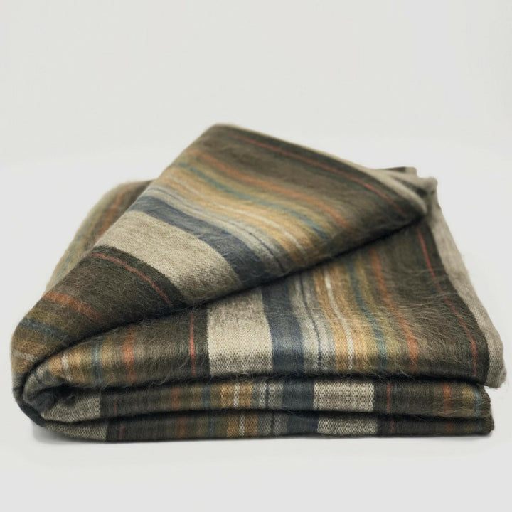 Atocha alpaca wool throw blanket – a folded, striped blanket featuring green, brown, blue, and beige hues, rests on a plain surface. The soft texture suggests warmth and comfort in a minimalist setting
