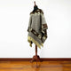A poncho is draped on a mannequin, featuring brown and beige geometric designs, with fringed edges, standing on a wooden floor against a plain white wall.