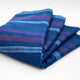 Tamboloma baby alpaca wool blanket – a folded blue blanket with horizontal stripes in shades of purple and pink, resting on a plain white surface