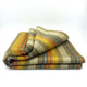 Rumi baby alpaca wool blanket – a folded, multicolored striped blanket lies on a surface. The pattern includes shades of orange, yellow, brown, and gray, set against a plain, white background