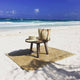 A straw hat and bag rest on a wooden stool atop a woven mat on a sandy beach, with a turquoise ocean and clear blue sky in the background.