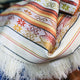 A woven fabric displays intricate geometric and floral patterns with red, yellow, and orange stripes. It features white fringe tassels, creating a decorative textile laid on a surface.
