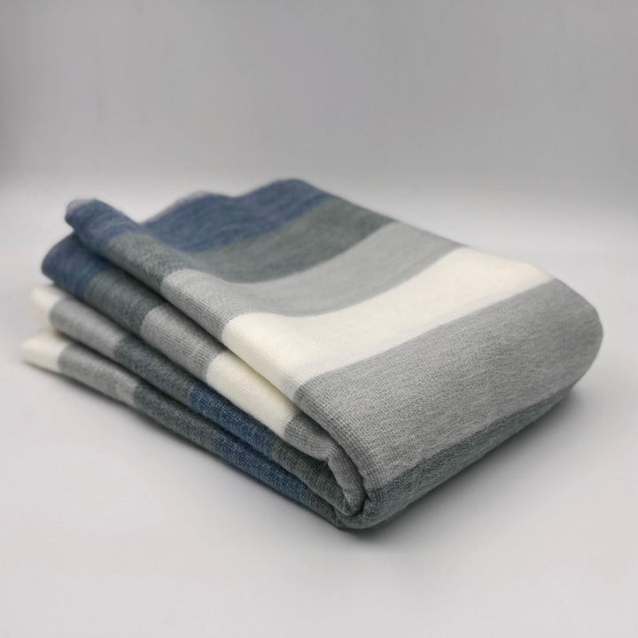Poguios baby alpaca wool blanket – a folded blanket with wide, horizontal stripes in shades of blue, gray, and white lies on a smooth, neutral-colored surface in a minimalistic setting