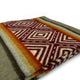 A folded blanket features a geometric design with red and white diamond patterns. It includes stripes in shades of gray, orange, and green on a smooth surface, suggesting warmth and style.