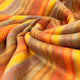 Cumanda baby alpaca wool blanket – a multicolored, striped fabric with warm tones is softly draped, creating gentle folds and ripples. The texture appears soft and woolly, suggesting a cozy, inviting material