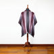 A decorative poncho with maroon and gray vertical stripes drapes on a mannequin stand, set against a plain white wall and wooden floor background.