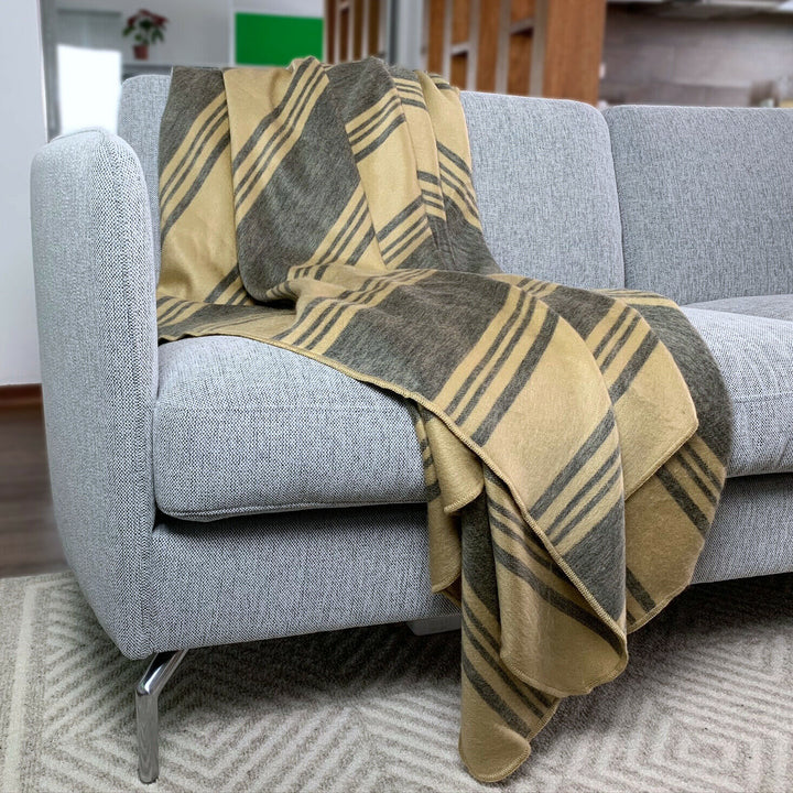 Chalua baby alpaca wool blanket – a beige and gray striped blanket drapes casually over a modern gray sofa in a living room with wooden elements and a patterned blanket