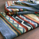 A folded, multicolored blanket with geometric patterns rests on a wooden surface, featuring shades of gray, red, orange, and green in a zigzag and striped design.