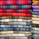 Wholesale lot of 10 queen alpaca wool blankets – stacks of folded, colorful, striped textiles rest neatly in horizontal layers, featuring vibrant reds, purples, blues, and earthy tones, creating a visually rich and orderly display