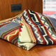 A folded, patterned blanket rests on a wooden surface. The design features geometric shapes in earthy tones. A power outlet is visible on the wooden wall in the background.