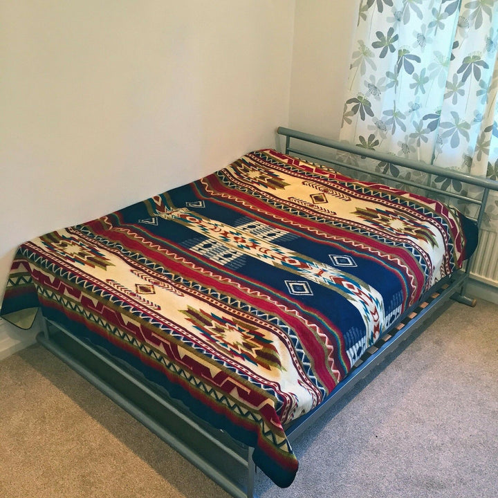 Metal bed frame holds a bed covered with a colorful geometric patterned blanket, situated in a bedroom with beige carpet and partially visible floral-patterned curtains.