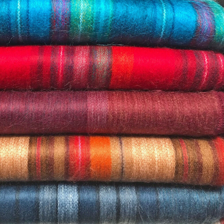 Wholesale lot of 15 queen alpaca wool blankets – colorful wool blankets are stacked neatly. Each blanket has a unique pattern of stripes in various colors, including blue, red, brown, and orange. The setting is a close-up view