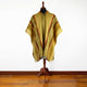 Poncho displayed on a mannequin, features earthy tones with geometric patterns. It hangs over a wooden stand against a plain white background, positioned on a polished wooden floor.