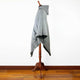 Gray woolen cloak hangs on a wooden mannequin stand, showing a draped design with a hood, and pointed hem, positioned against a plain white wall with a wooden floor.