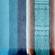 A woolen fabric features vertical stripes in varying shades of blue, gray, and red, with a tribal pattern on the right and fringe at the bottom edge.