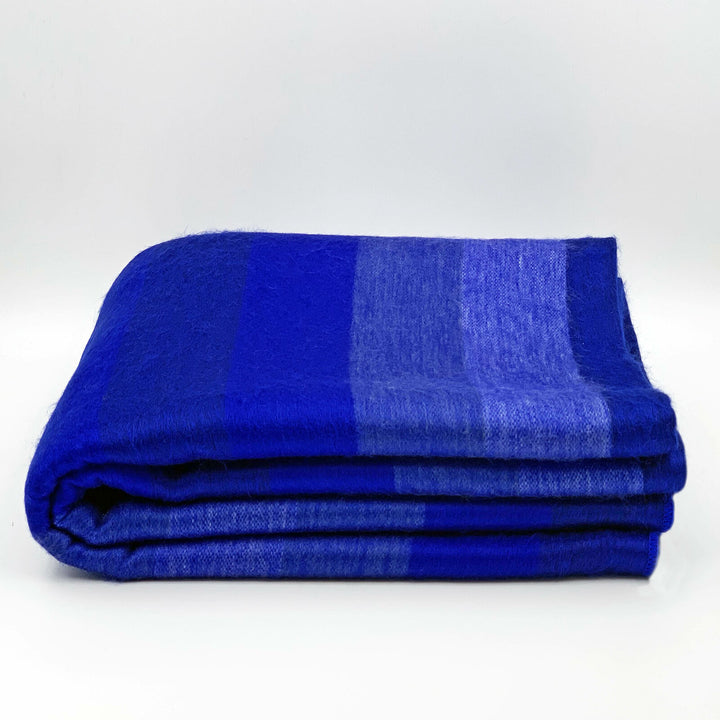 Chibuleo baby alpaca wool blanket – a folded blue blanket features alternating dark and light gradient stripes, resting on a clean, white background. No text is present