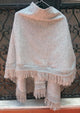 A light gray shawl with fringed edges hangs on a hanger. It is draped in front of a decorative black metal gate, providing a textured backdrop.