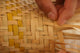Hands weave thin bamboo strips into a textured, basket-like pattern, incorporating light yellow strands. The setting suggests a manual crafting process focused on intricate detail.
