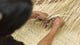 Hands weaving dried palm fronds together on a woven mat, creating a basket in an outdoor setting. The fronds crisscross, forming a textured pattern.