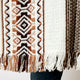 A woven textile features geometric patterns in brown and white, with fringes at the bottom, draped over a person wearing dark pants.