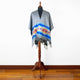 A grey poncho with a vibrant geometric pattern and fringes drapes over a mannequin on a wooden stand, set against a white wall and wooden floor.