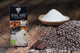 A box of PACARI Premium Organic Chocolate Cuzco Pink Salt & Nibs stands on a wooden surface beside a mound of cocoa nibs, chocolate squares, and a bowl of coarse salt.