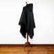 A black fringed poncho with a hood drapes over a wooden mannequin stand, set against a plain white wall and wooden floor.
