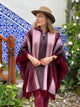 A person wearing a striped poncho and hat stands outdoors, looking to the side. The background features decorative blue and white tiles, wooden railing, and blooming flowers.