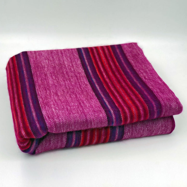 Cayetana baby alpaca wool blanket – a folded, striped pink and purple blanket lies neatly on a plain white surface. It features alternating horizontal bands of vibrant colors, creating a cozy, inviting appearance