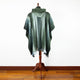 A green, hooded poncho is draped over a wooden mannequin stand, displayed against a white wall and polished wooden floor, highlighting its draped, flowing design.