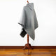 A gray, hooded poncho drapes over a wooden mannequin stand, set against a white background and wooden floor. No text is present.