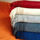 Stacked blankets with fringed edges rest in a gradient from red to blue on an orange textured surface, showcasing a cozy textile arrangement.