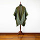 A green poncho with white and red stripes is displayed on a wooden stand, set against a plain white wall and wooden floor.