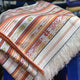 A woven textile displays colorful stripes and intricate patterns, including geometric shapes and stylized birds, draped over a surface with long fringes, creating a vibrant and textured appearance.